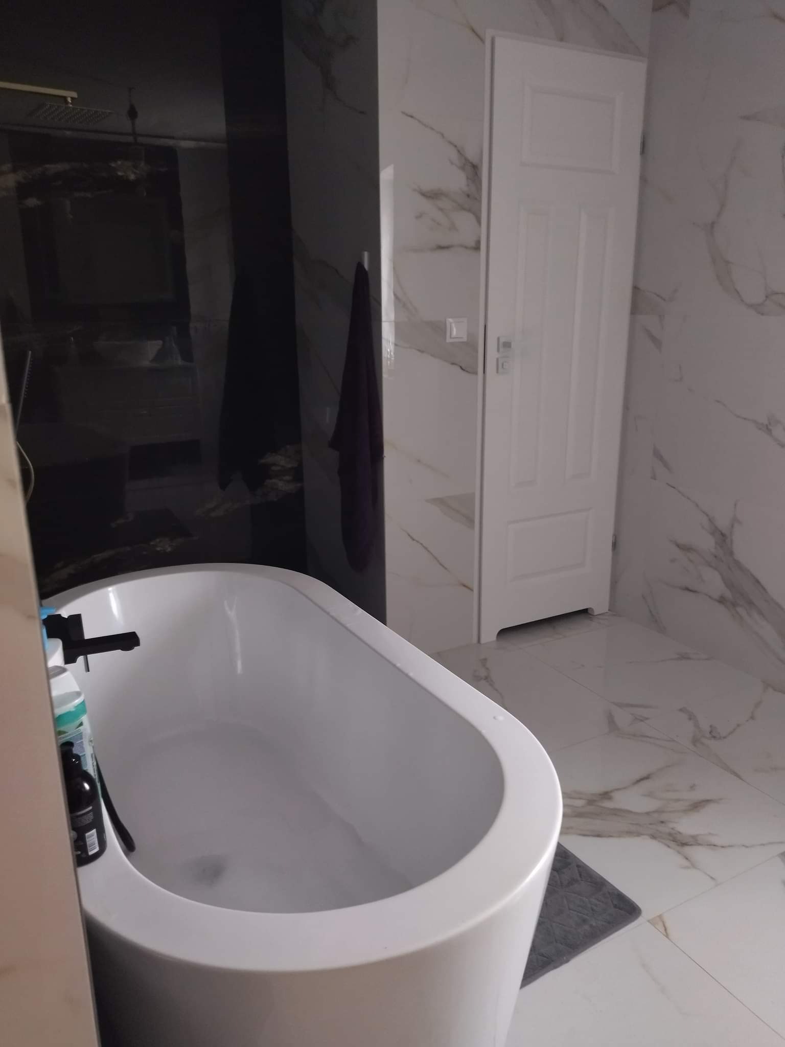 Nowoczesna łazienka z wolnostojącą białą wanną, marmurowymi płytkami na ścianach i podłodze oraz czarną kabiną prysznicową z deszczownicą.