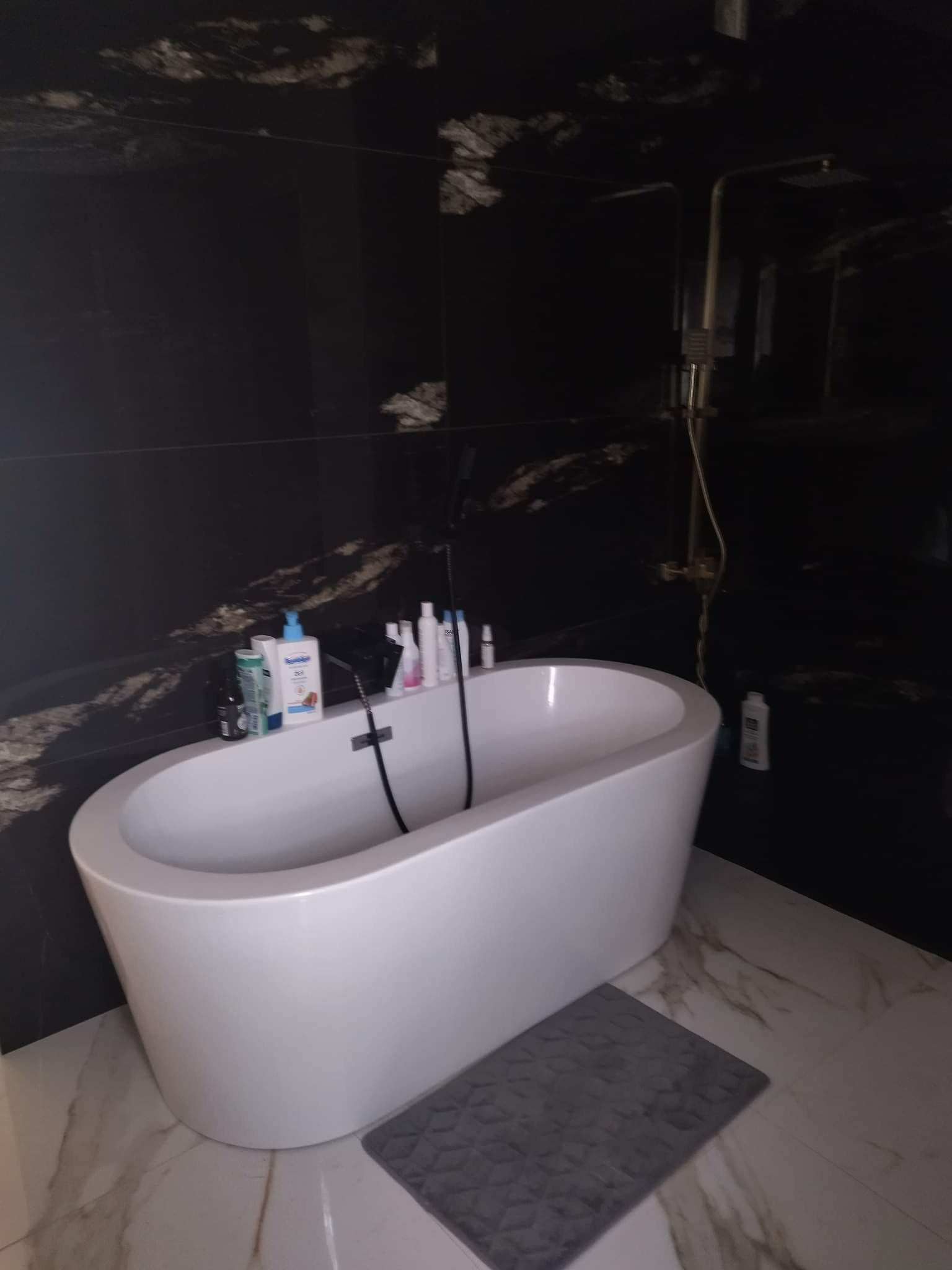 Nowoczesna łazienka z wolnostojącą białą wanną na tle czarnych płytek imitujących marmur, szara wycieraczka, złota bateria prysznicowa, kosmetyki na rancie wanny.