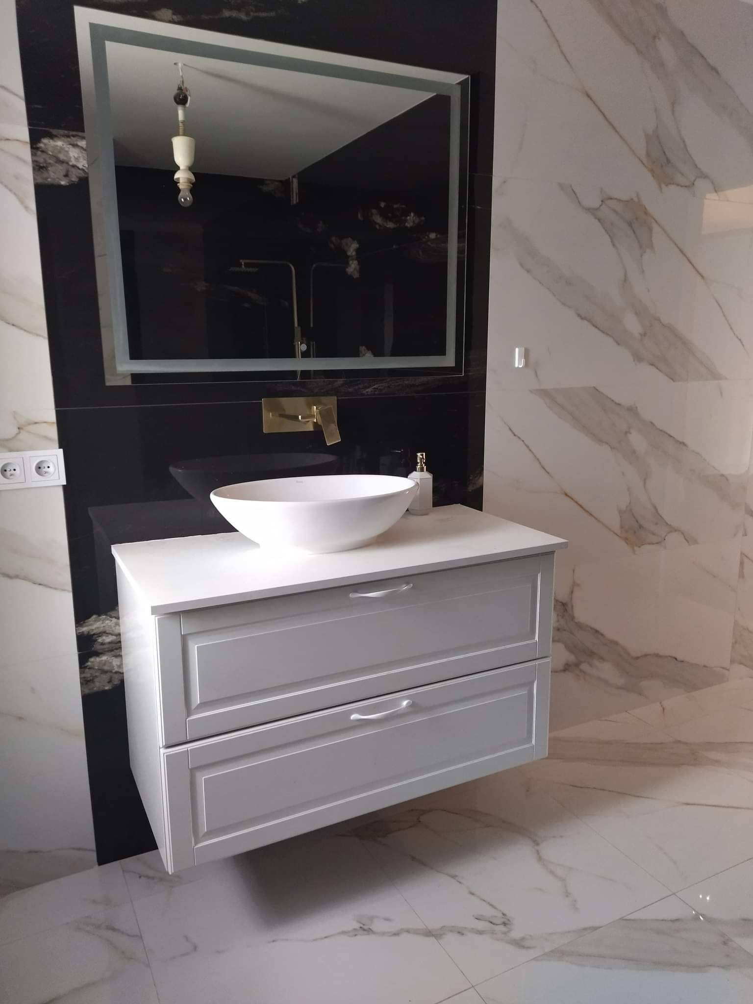 Nowoczesna łazienka z umywalką nablatową w kształcie misy, białą szafką pod umywalką, lustrem z podświetleniem i marmurowymi płytkami na ścianach i podłodze.