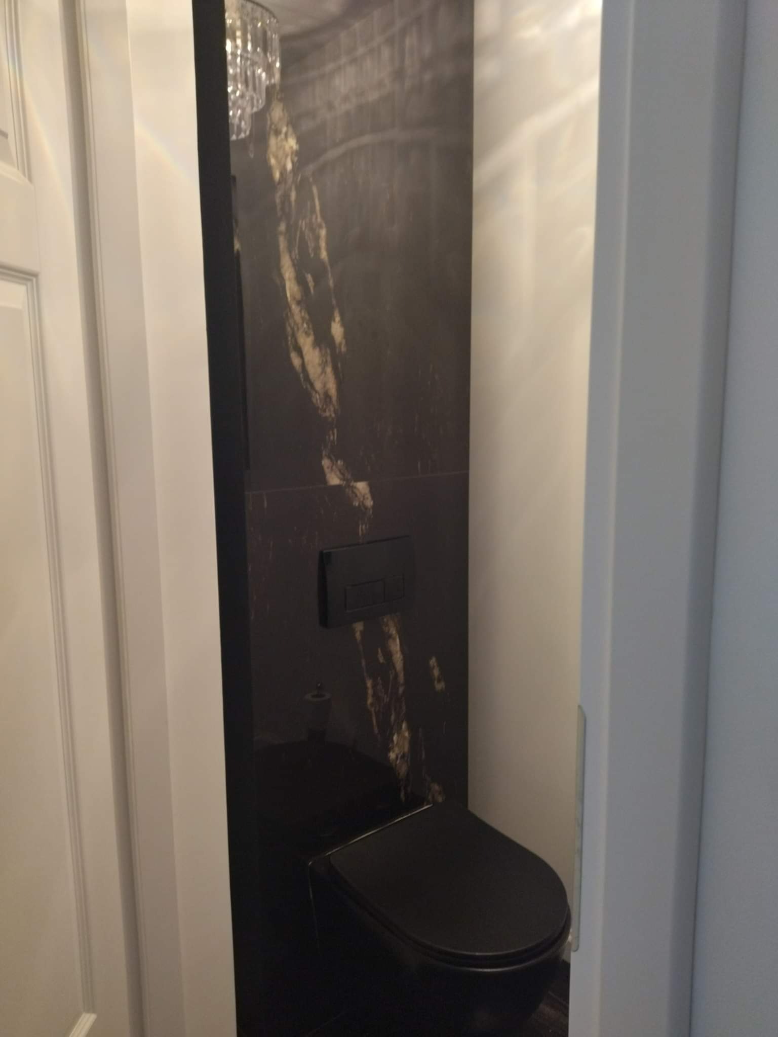 Ciemna toaleta z czarną miską WC i płytkami imitującymi marmur, oświetlona kryształowym żyrandolem, widoczna zza białej ościeżnicy drzwi.