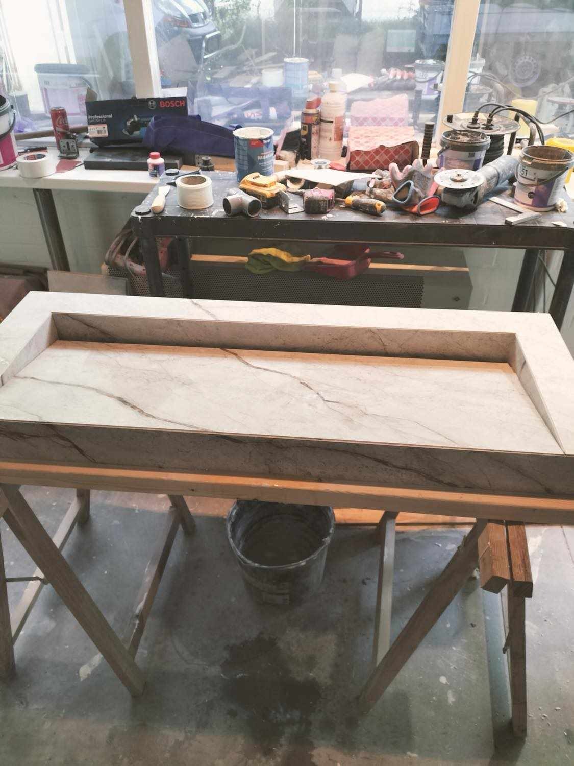 Blat z jasnego marmuru z ciemniejszymi żyłami, osadzony w drewnianej ramie na kozłach, w tle warsztatowy nieład z narzędziami i materiałami budowlanymi.