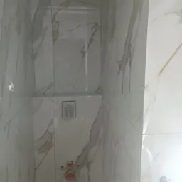 Ściana łazienki wyłożona białymi płytkami imitującymi marmur z widocznymi przyłączami wodno-kanalizacyjnymi i panelem sterującym podtynkowym.