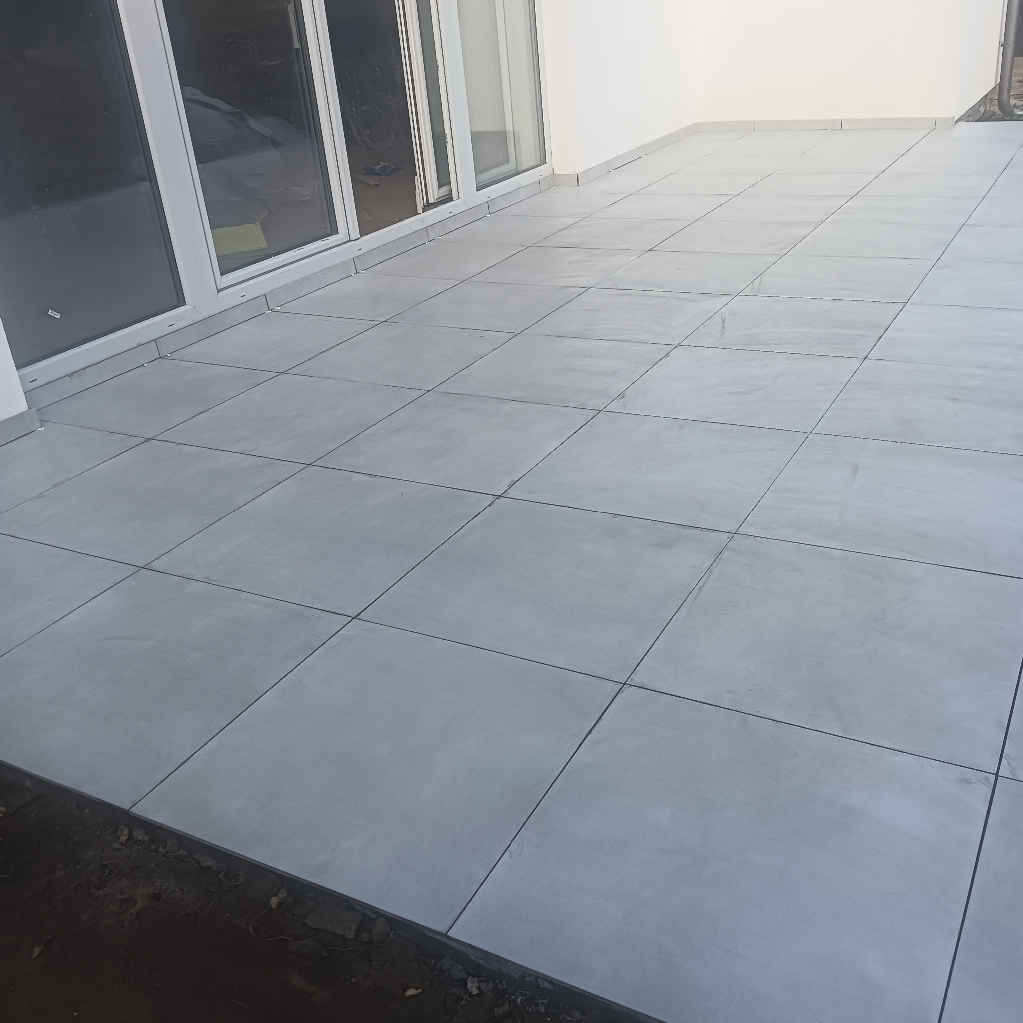 Wyłożony jasnoszarą, kwadratową glazurą taras z widocznymi szklanymi drzwiami przesuwnymi i fragmentem ziemi wzdłuż krawędzi.