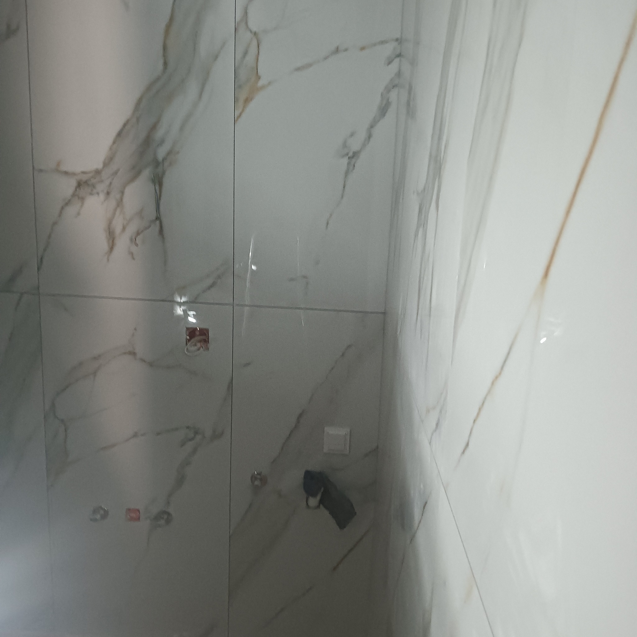 Ściana łazienki wyłożona białymi płytkami imitującymi marmur z widocznymi puszkami instalacyjnymi i uchwytem na ręcznik z zawieszonym ciemnym ręcznikiem.