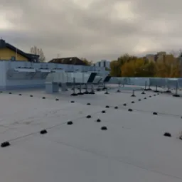 Szare kanały wentylacyjne na płaskim dachu budynku, widoczne czarne punkty mocowania instalacji, tło stanowi zabudowa miejska i pochmurne niebo.