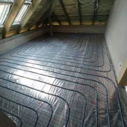 Niebieskie rury ogrzewania podłogowego ułożone na folii refleksyjnej z czerwoną siatką w surowym stanie, widok z góry pod skosem dachu z oknami.