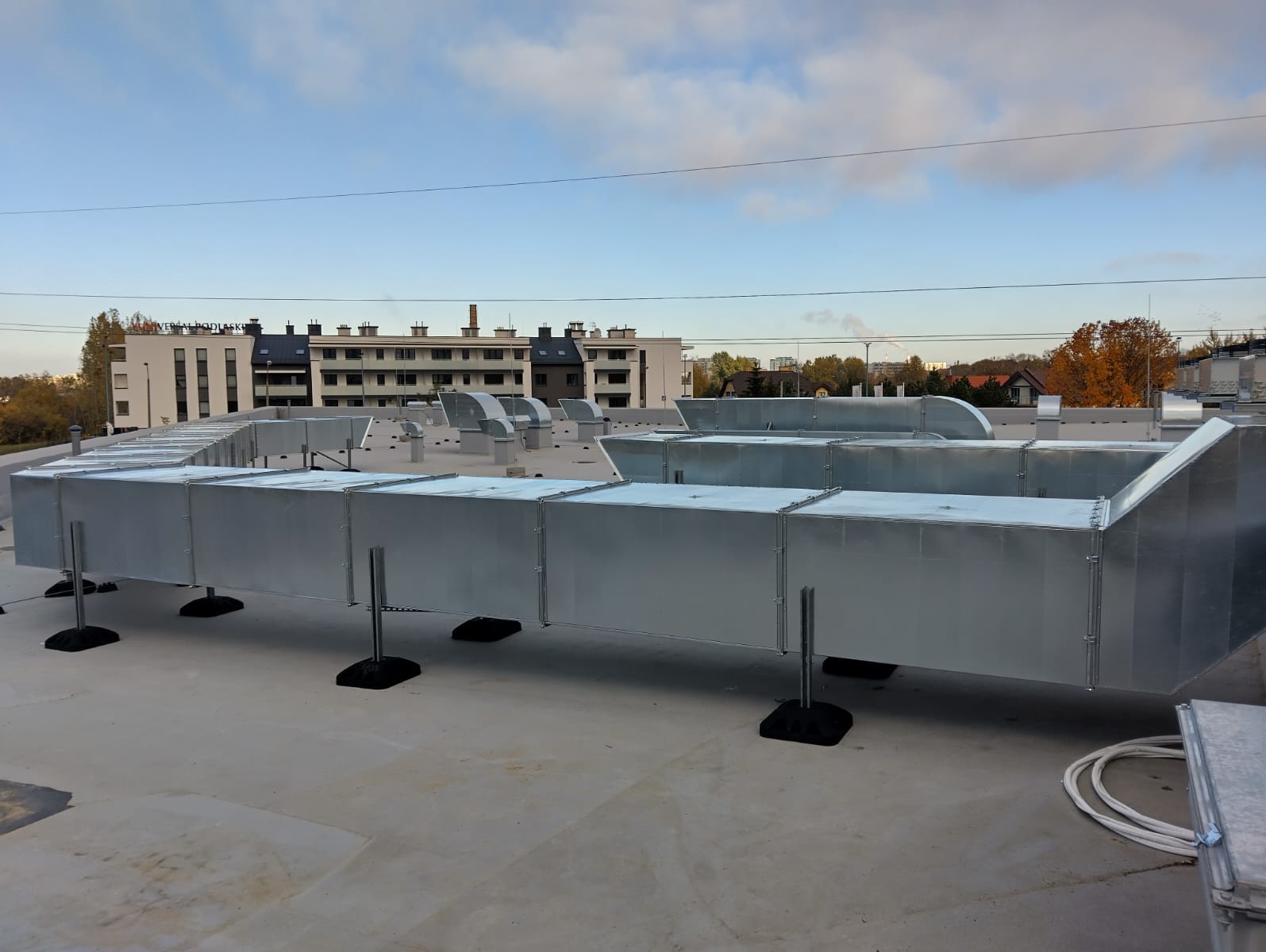 System kanałów wentylacyjnych z blachy ocynkowanej, zamontowany na płaskim dachu budynku, z widocznymi elementami mocującymi i tłem zabudowy miejskiej pod błękitnym niebem.