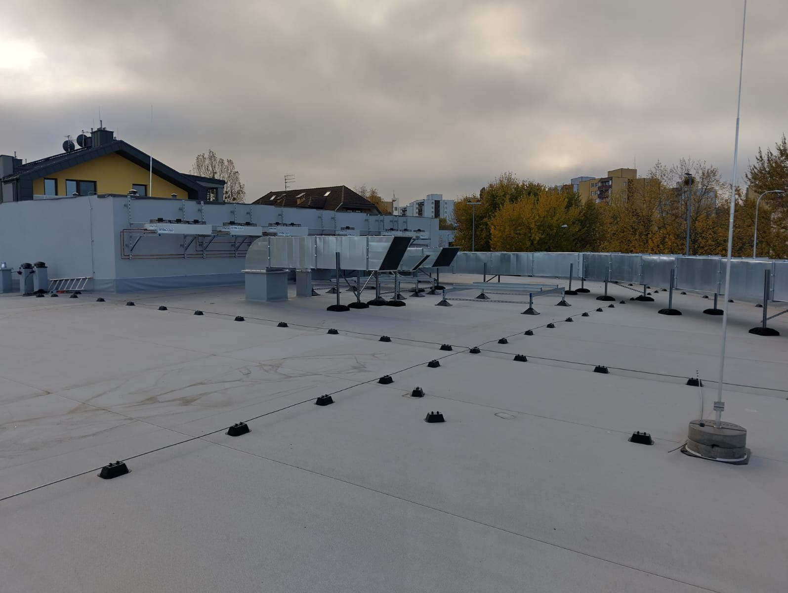 Szare kanały wentylacyjne na płaskim dachu budynku, widoczne czarne punkty mocowania instalacji, tło stanowi zabudowa miejska i pochmurne niebo.