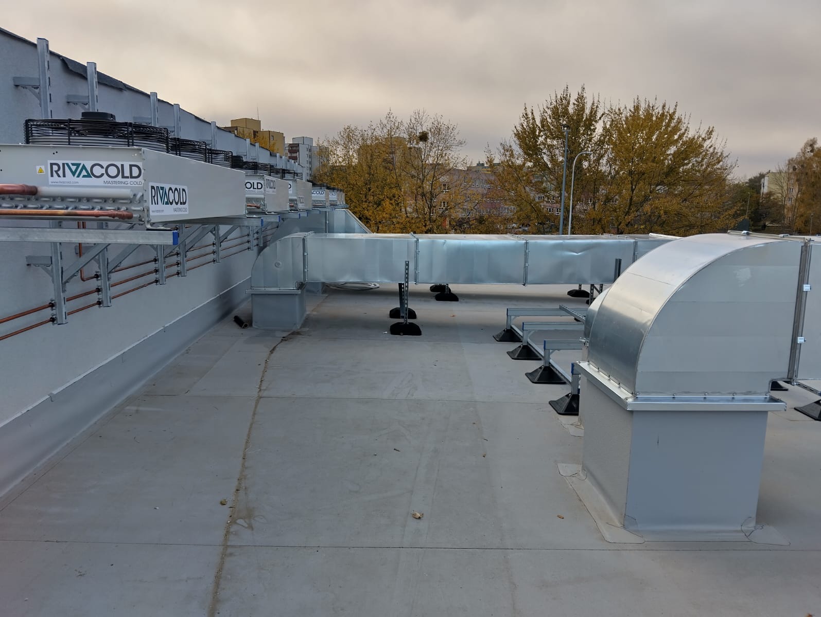 Instalacja kanałów wentylacyjnych na dachu budynku, widoczne elementy systemu i jednostki zewnętrzne RIVACOLD, jesienne drzewa w tle.