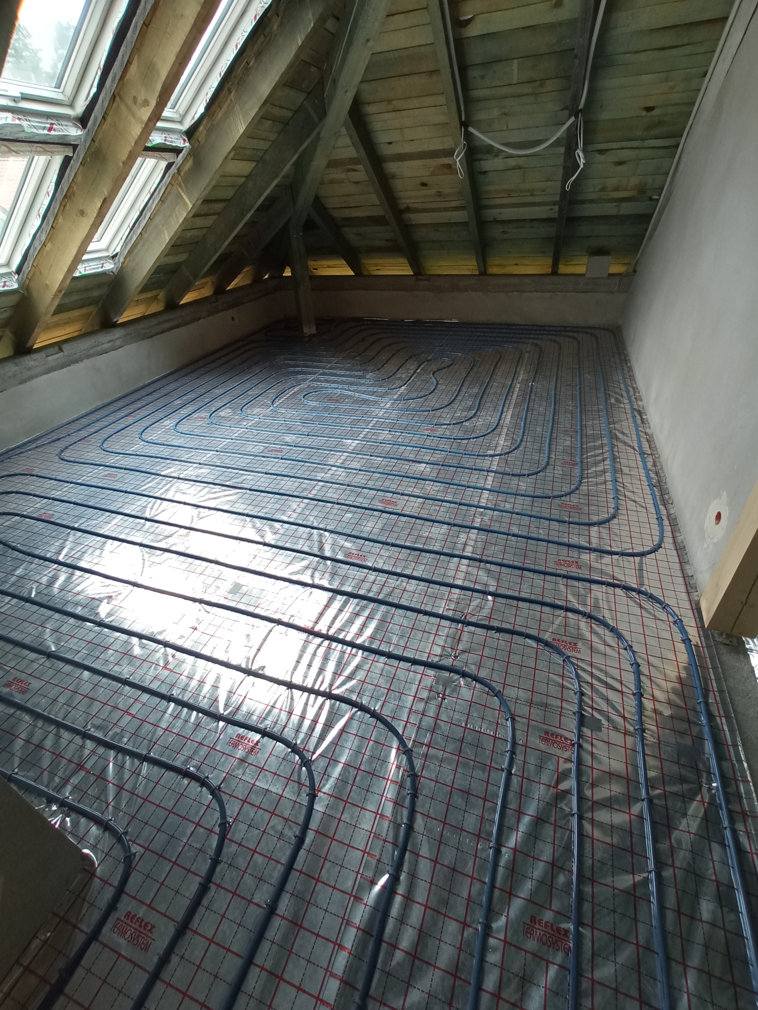 Niebieskie rury ogrzewania podłogowego ułożone na folii refleksyjnej z czerwoną siatką w surowym stanie, widok z góry pod skosem dachu z oknami.