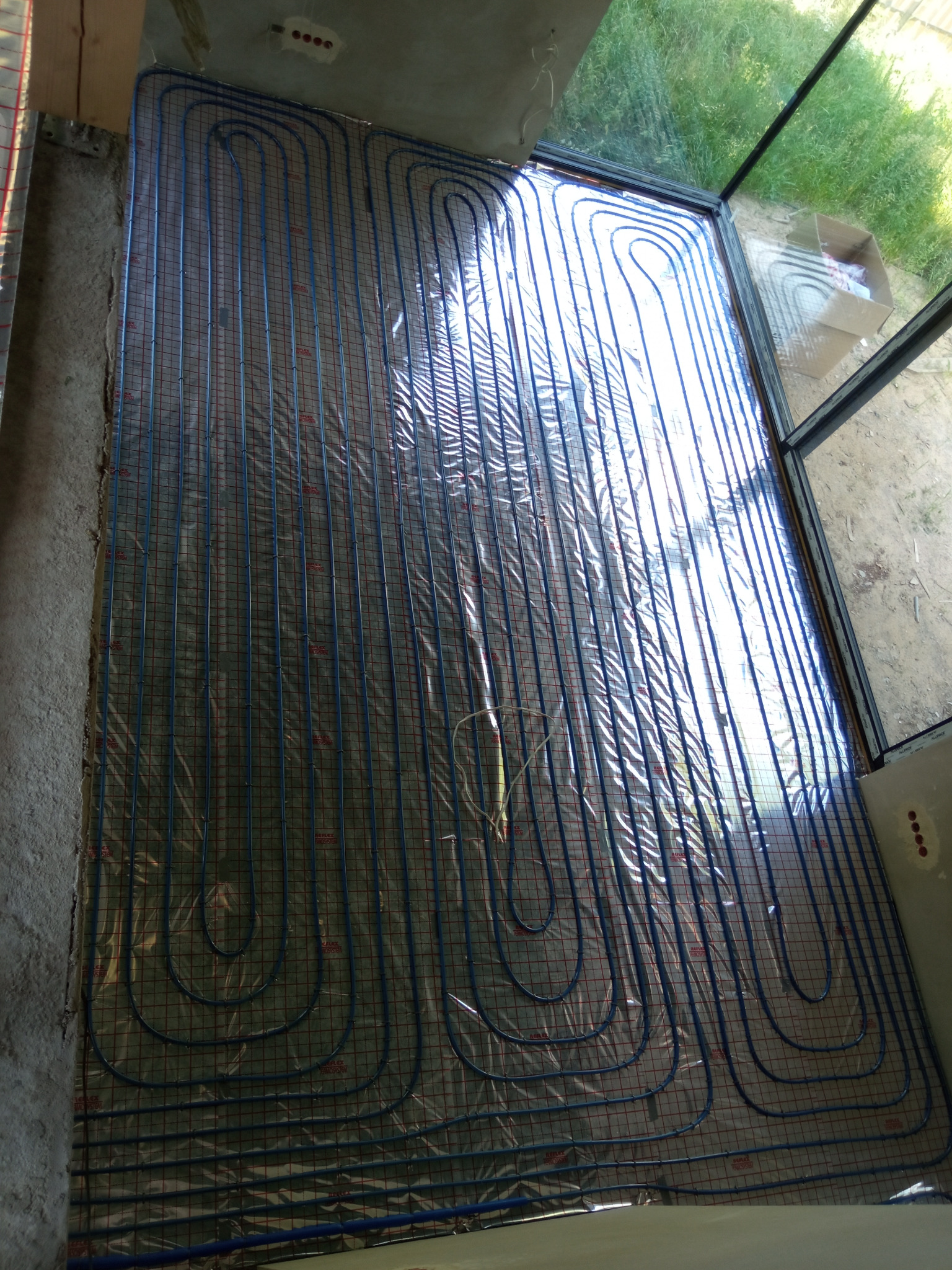 Niebieskie rurki ogrzewania podłogowego ułożone na folii z widoczną siatką, przygotowane do zalania wylewką, widok z góry na surowy stan wykończenia pomieszczenia.