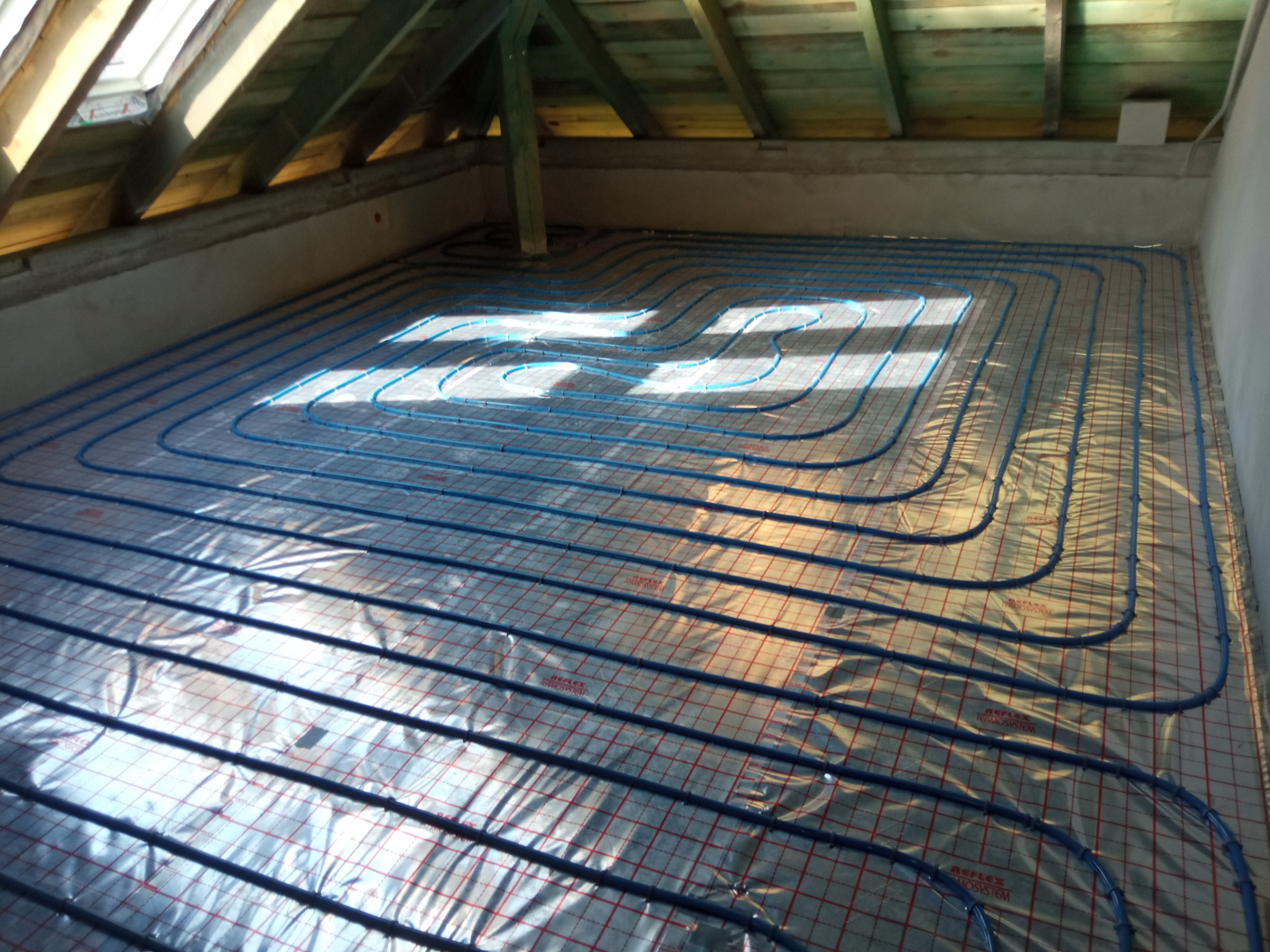 Niebieskie rury ogrzewania podłogowego ułożone na folii refleksyjnej z czerwoną siatką, widok z góry w surowym stanie poddasza z drewnianą konstrukcją dachu i oknem dachowym.