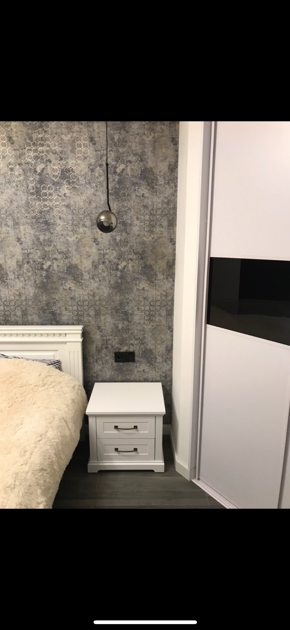 Sypialnia z wzorzystą tapetą w odcieniach szarości, wiszącą lampą z metalową kulą, białym stolikiem nocnym z dwoma szufladami i częścią białej szafy przesuwnej z czarnym pasem.