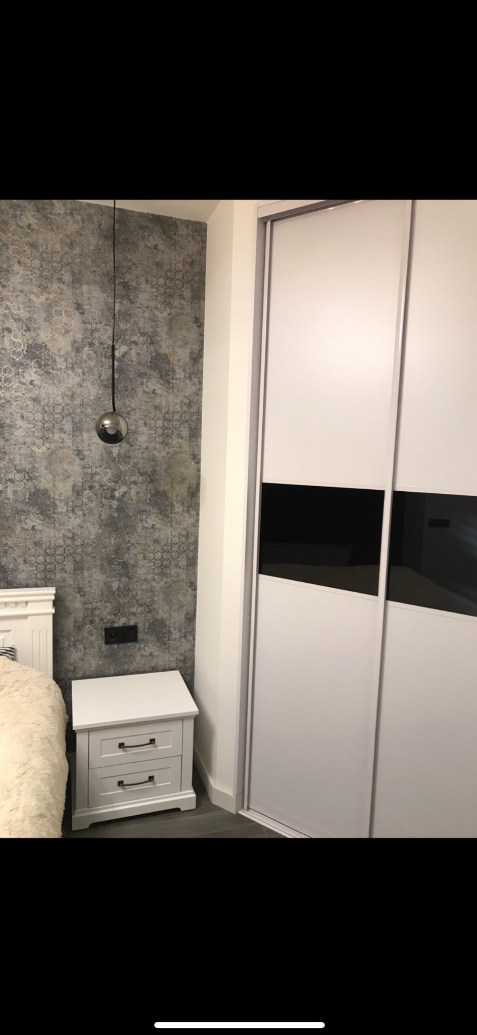 Sypialnia z szarą, wzorzystą tapetą, kulistą lampą wiszącą, białą szafką nocną z dwiema szufladami i białą szafą przesuwną z czarnymi akcentami.