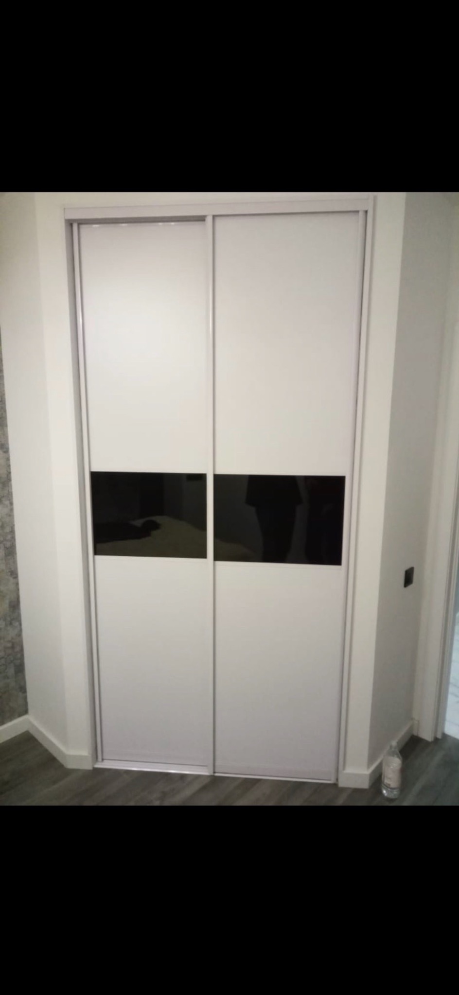 Biała szafa przesuwna z czarnymi, prostokątnymi wstawkami w dolnej części, widoczna w rogu pomieszczenia z szarą podłogą i butelką wody przy ścianie.