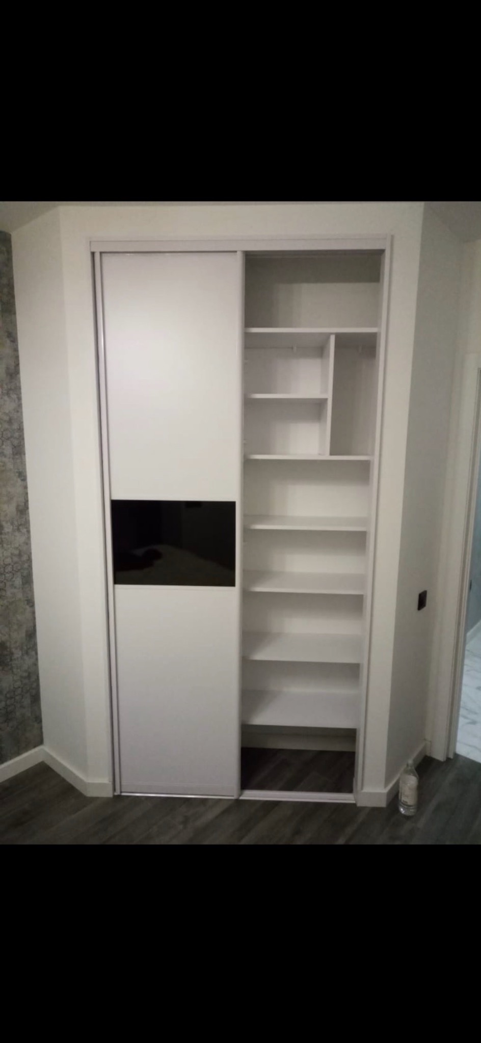 Biała szafa wnękowa z przesuwnymi drzwiami, częściowo otwartymi, ukazująca puste półki, z czarnym prostokątnym panelem na drzwiach, na tle szarej ściany i podłogi z paneli.