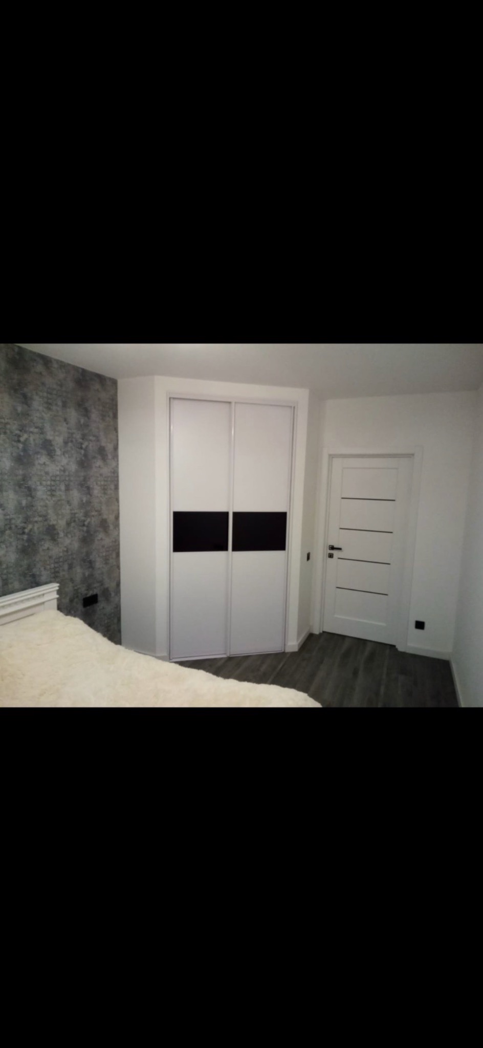 Biała szafa przesuwna z czarnymi akcentami w sypialni z szarą tapetą i drewnianą podłogą, widoczny fragment białego łóżka.