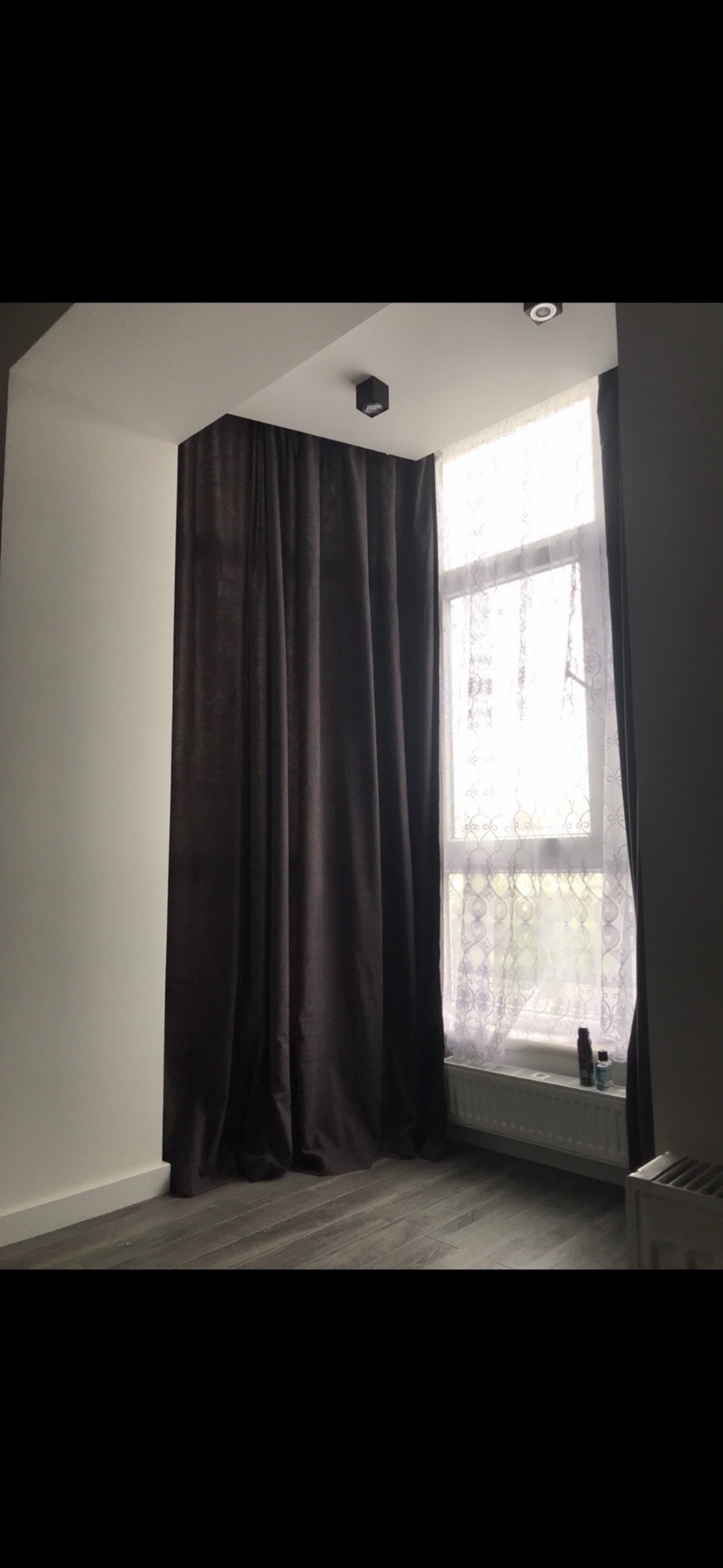 Pokój z szarymi panelami podłogowymi, białą ścianą i oknem z brązowymi zasłonami oraz białą firanką z delikatnym wzorem, oświetlony kwadratowym plafonem.