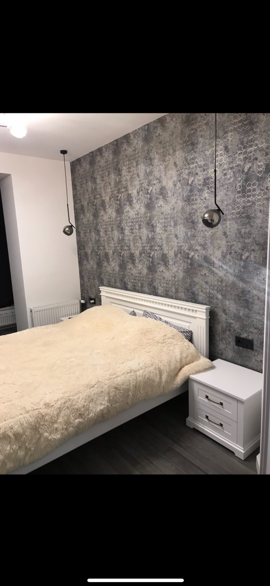 Sypialnia z łóżkiem z białym zagłówkiem, puszystą beżową narzutą, białą szafką nocną i tapetą w geometryczne wzory w odcieniach szarości, oświetlona wiszącymi lampami z metalowymi kulami.