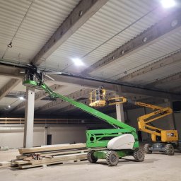 EL-ELEKTRO Łukasz Gaborski - Montaż oświetlenia w hali przemysłowej z użyciem podnośników koszowych. Widoczne elementy instalacji i pracownicy na wysokościach.