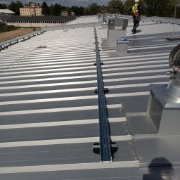 EL-ELEKTRO Łukasz Gaborski - Montaż instalacji odgromowej na dachu z blachy trapezowej. Widoczny pracownik w kasku i uprzęży. Szyna montażowa na dachu, wentylatory dachowe. Budynek w tle.