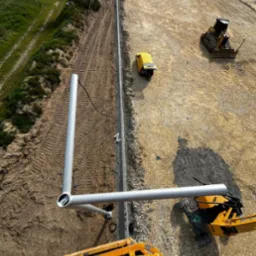 Montaż słupa oświetleniowego z podnośnika koszowego na terenie budowy, widok z góry, w tle budynek i sprzęt budowlany.