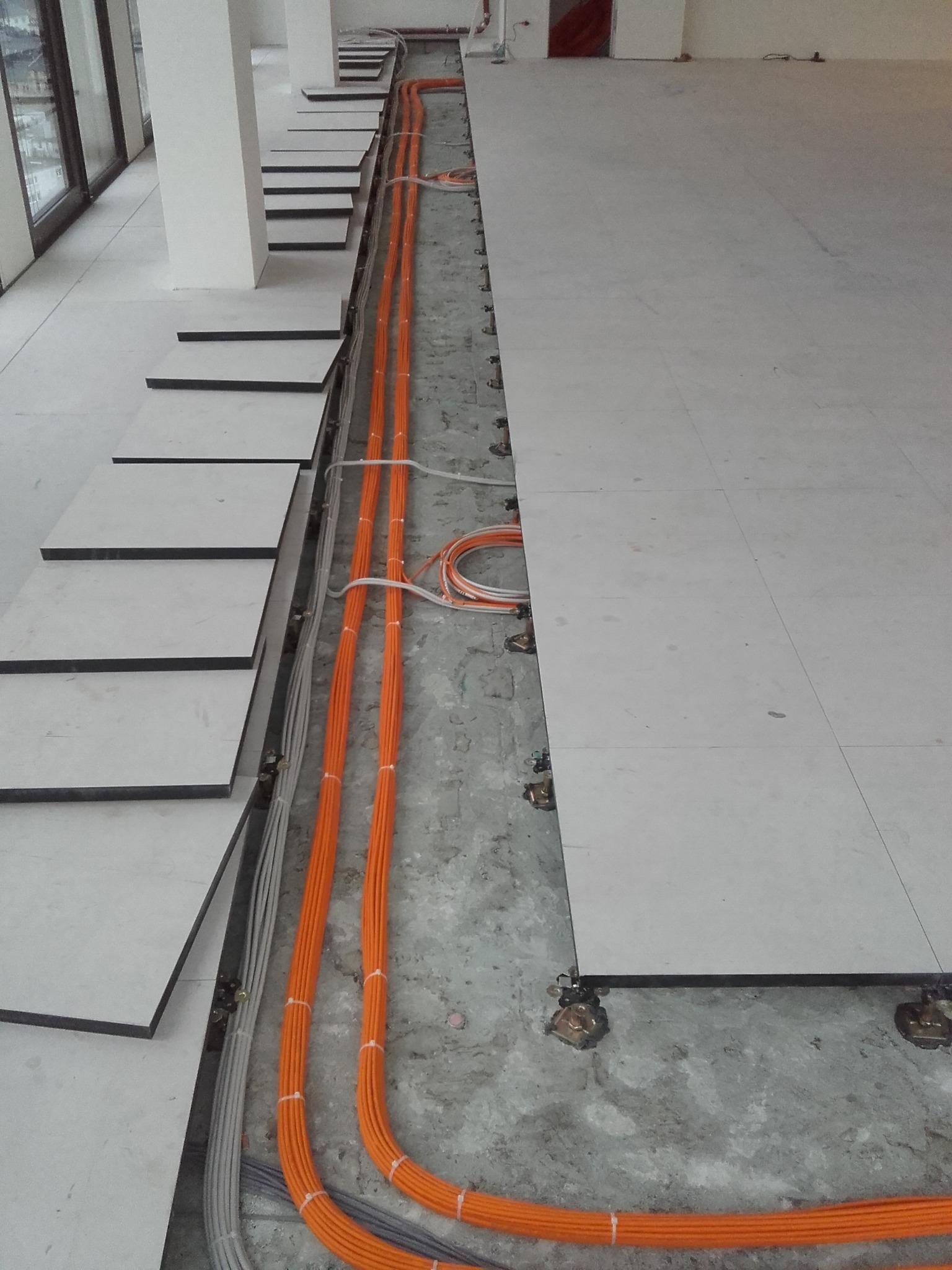 Pomarańczowe i szare kable ułożone pod podniesioną podłogą z jasnych płyt. Widoczne elementy mocujące podłogę. Część podłogi jest jeszcze nieułożona.