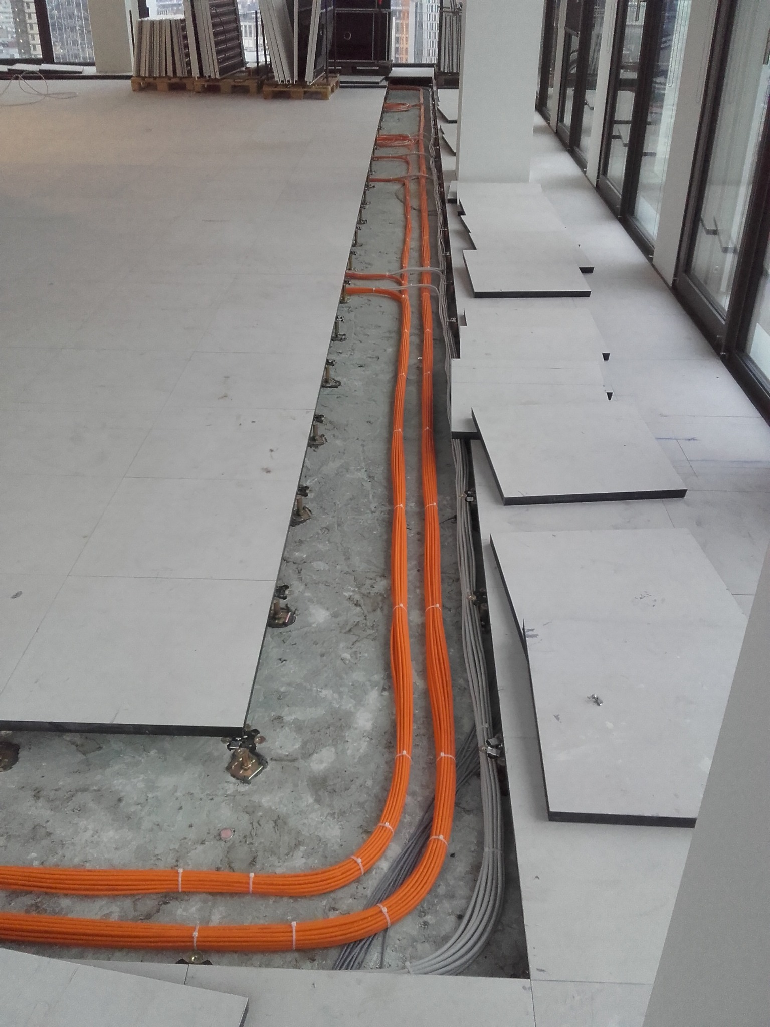 Montaż podłogi podniesionej z widocznymi pomarańczowymi kablami instalacji elektrycznej w przestrzeni biurowej, z oknami w tle.
