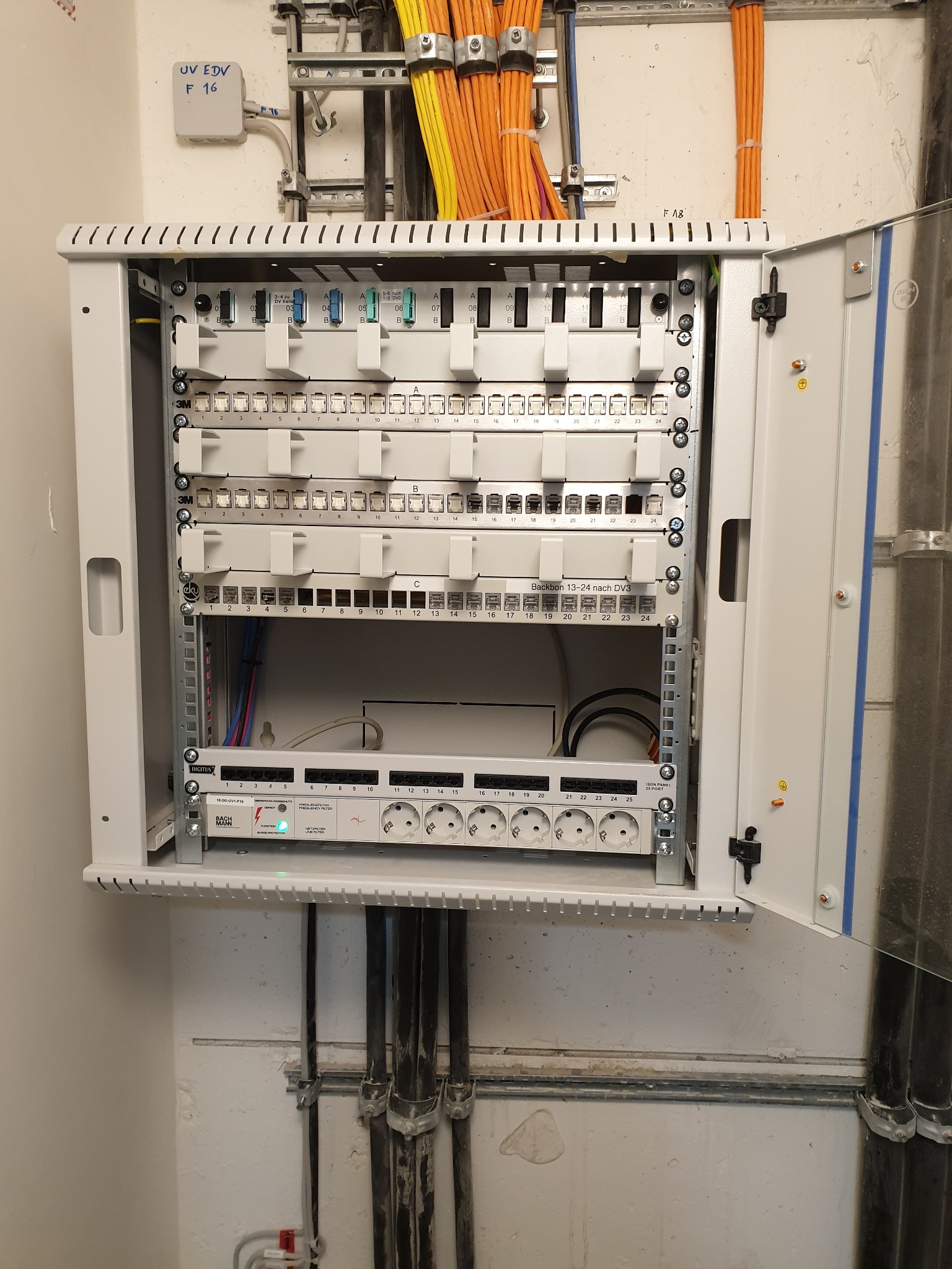 Otwarta szafa rack z patch panelem, gniazdami zasilającymi i okablowaniem w kolorach żółtym i pomarańczowym. Widoczne oznaczenia portów i wskaźnik LED.