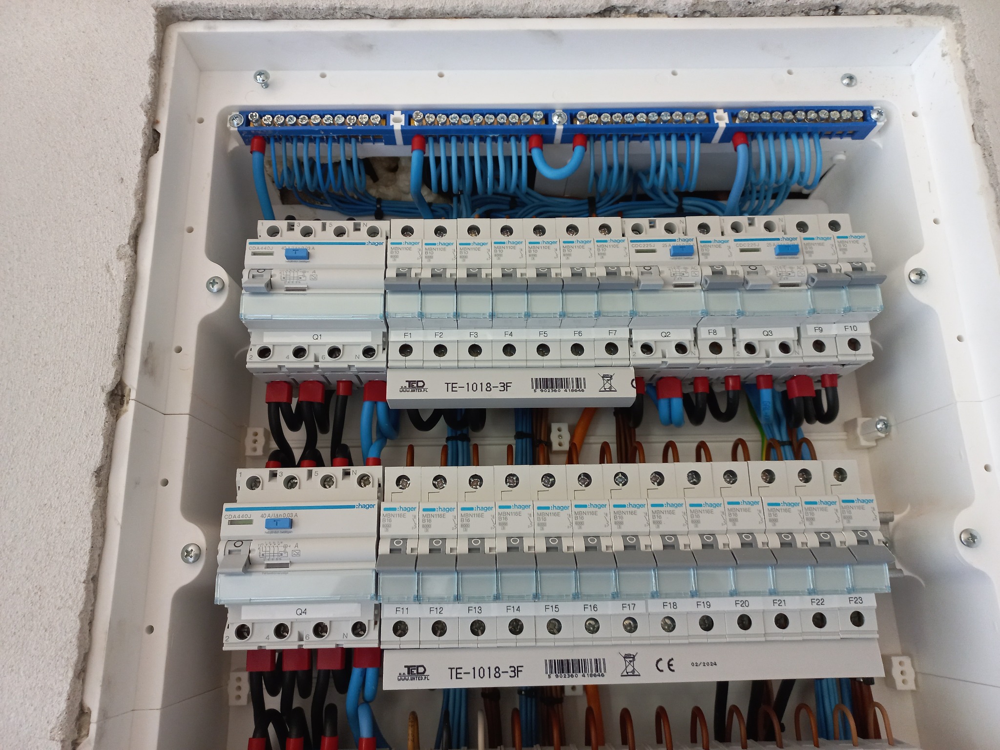 Rozdzielnica elektryczna Hager z bezpiecznikami i okablowaniem wewnątrz białej obudowy, widoczne oznaczenia TE-1018-3F i terminale z niebieskimi przewodami.