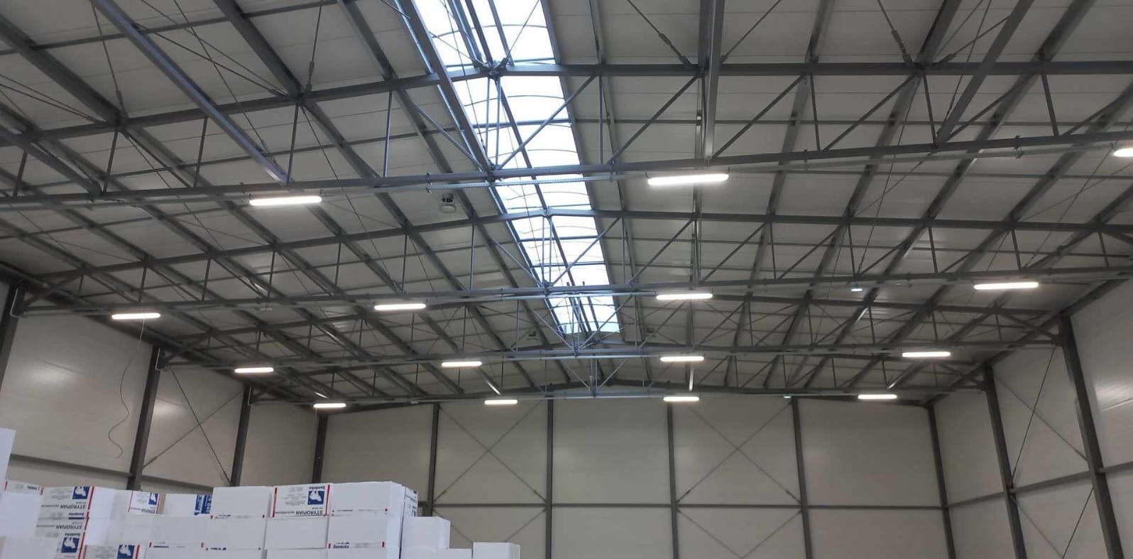 Oświetlenie hali magazynowej z widoczną metalową konstrukcją dachu i świetlikami. Lampy LED równomiernie rozmieszczone. Białe ściany, w dole palety ze styropianem.