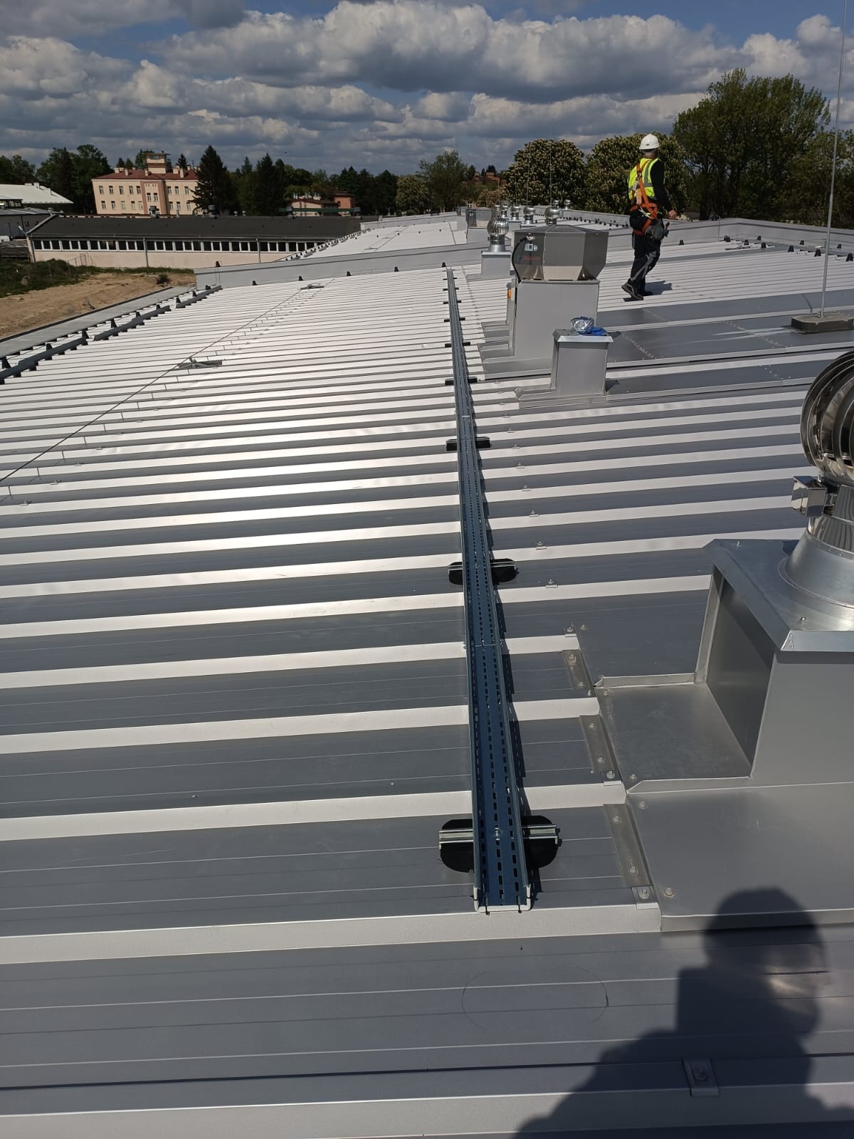 Montaż instalacji odgromowej na dachu z blachy trapezowej. Widoczny pracownik w kasku i uprzęży. Szyna montażowa na dachu, wentylatory dachowe. Budynek w tle.