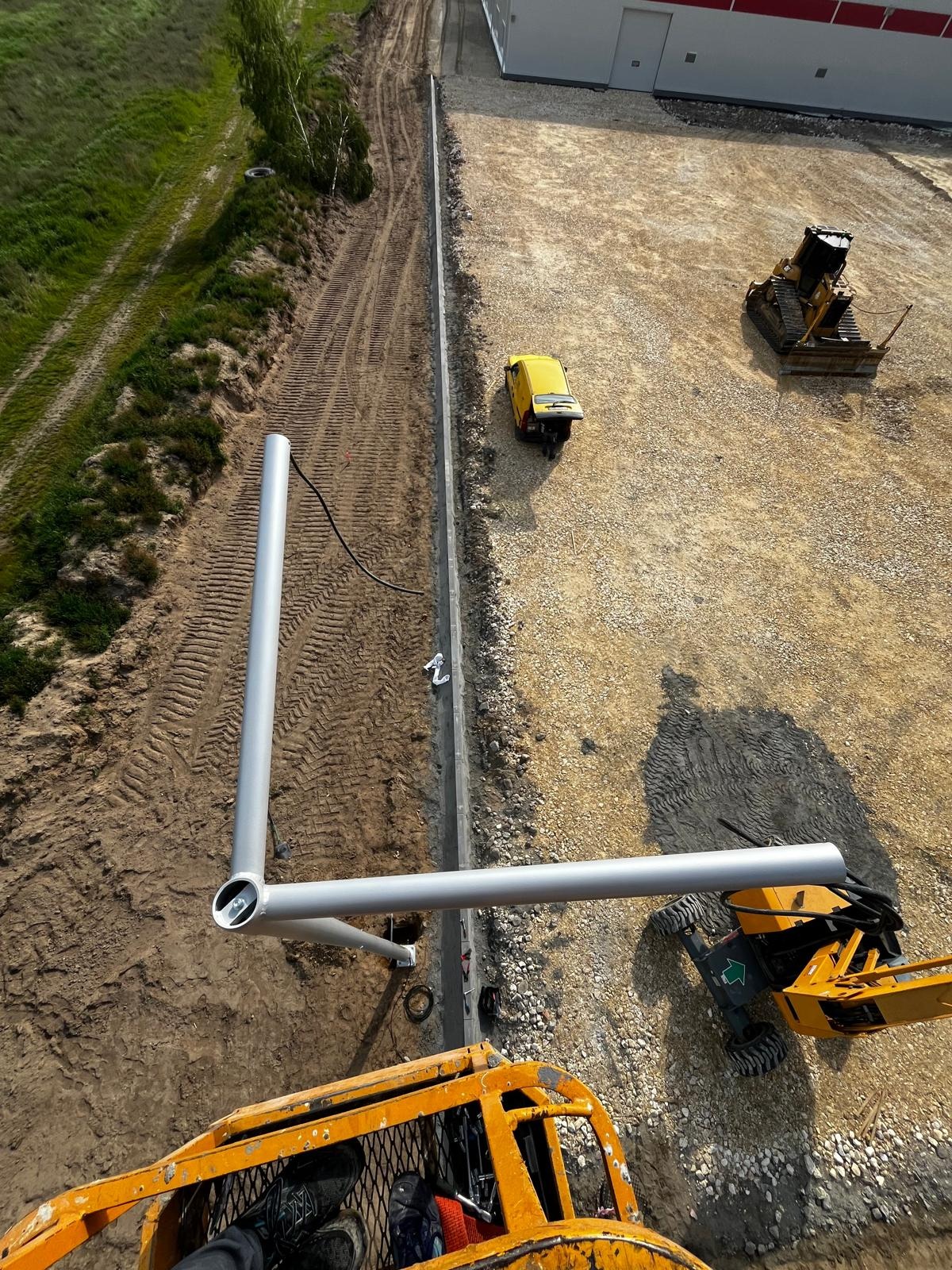 Montaż słupa oświetleniowego z podnośnika koszowego na terenie budowy, widok z góry, w tle budynek i sprzęt budowlany.