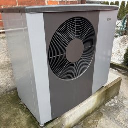 HYDROLINE SPÓŁKA Z OGRANICZONĄ ODPOWIEDZIALNOŚCIĄ - Szaro-grafitowa pompa ciepła na betonowym fundamencie, z widoczną ceglaną ścianą w tle i elementami zieleni. Widoczne ślady wilgoci na urządzeniu i podłożu.