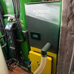 HYDROLINE SPÓŁKA Z OGRANICZONĄ ODPOWIEDZIALNOŚCIĄ - Szaro-żółty piec Pereko Qm Pell EVO 8kW w kotłowni, widoczne rury instalacji grzewczej i sterownik. Ściany pomalowane na zielono, podłoga wyłożona płytkami.