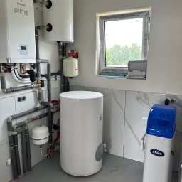 Nowoczesna kotłownia z piecem gazowym Galmet Prima, zbiornikami i zmiękczaczem wody Unico, w jasnym pomieszczeniu z oknem. Instalacja po montażu, widoczne rury i podłączenia.