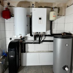 Kompletna instalacja grzewcza w kotłowni: piec gazowy Nibe, zbiorniki buforowe (czerwony i biały), szary zasobnik CWU i czarny zmiękczacz wody Unico. Białe płytki na ścianach.