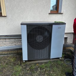 HYDROLINE SPÓŁKA Z OGRANICZONĄ ODPOWIEDZIALNOŚCIĄ - Szara pompa ciepła Nibe F2120 zamontowana przy ścianie budynku z dwoma oknami. Widoczne rury instalacyjne i fragment trawnika. Na urządzeniu leży zielona ściereczka.