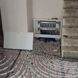 HYDROLINE SPÓŁKA Z OGRANICZONĄ ODPOWIEDZIALNOŚCIĄ - Rozłożone rury ogrzewania podłogowego na siatce, widoczny rozdzielacz w białej obudowie, surowe ściany i schody w tle. Instalacja w trakcie realizacji.