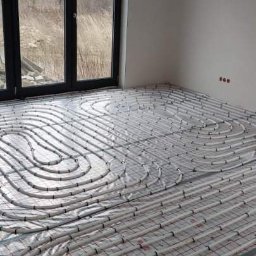 HYDROLINE SPÓŁKA Z OGRANICZONĄ ODPOWIEDZIALNOŚCIĄ - Białe rury ogrzewania podłogowego ułożone spiralnie na folii z czerwoną siatką w jasnym pomieszczeniu z dużym oknem. Widok z góry na instalację przed zalaniem posadzki.