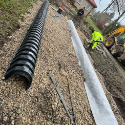 HYDROLINE SPÓŁKA Z OGRANICZONĄ ODPOWIEDZIALNOŚCIĄ - Instalacja drenażu rozsączającego: czarne rury drenażowe, geowłóknina i żwir. W tle pracownik w odblaskowej kamizelce i koparka. Prace ziemne przy domu.