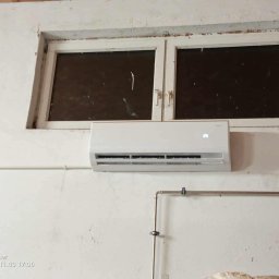 HYDROLINE SPÓŁKA Z OGRANICZONĄ ODPOWIEDZIALNOŚCIĄ - Biały klimatyzator ścienny zamontowany pod oknem na białej ścianie. Widoczne rury instalacji. Ujęcie z dołu, lekko z prawej strony.