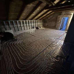 HYDROLINE SPÓŁKA Z OGRANICZONĄ ODPOWIEDZIALNOŚCIĄ - Instalacja ogrzewania podłogowego w poddaszu: rury ułożone na folii, widoczne elementy konstrukcji dachu i ścian. Prace instalacyjne w toku, widoczni pracownicy.