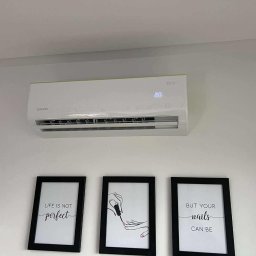 HYDROLINE SPÓŁKA Z OGRANICZONĄ ODPOWIEDZIALNOŚCIĄ - Biały klimatyzator Rotenso na ścianie nad trzema czarnymi ramkami z grafikami w salonie kosmetycznym. Widoczny wyświetlacz temperatury.