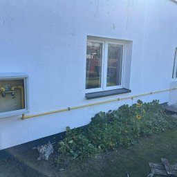 HYDROLINE SPÓŁKA Z OGRANICZONĄ ODPOWIEDZIALNOŚCIĄ - Biała ściana budynku z oknami i żółtą instalacją gazową, obok skrzynka gazowa. W tle zieleń i trawa. U dołu widoczna figurka psa.