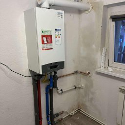 HYDROLINE SPÓŁKA Z OGRANICZONĄ ODPOWIEDZIALNOŚCIĄ - Biały, wiszący piec gazowy Ariston z podłączonymi rurami w kolorach: szarym, czerwonym i niebieskim. Widoczne fragmenty instalacji grzewczej w rogu pomieszczenia.