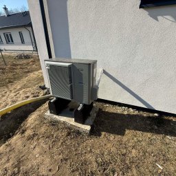 HYDROLINE SPÓŁKA Z OGRANICZONĄ ODPOWIEDZIALNOŚCIĄ - Szara pompa ciepła zamontowana na betonowej podstawie przy ścianie domu. Żółty przewód biegnie wzdłuż ziemi. Widoczny fragment budynku w tle.