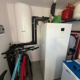 HYDROLINE SPÓŁKA Z OGRANICZONĄ ODPOWIEDZIALNOŚCIĄ - Biała pompa ciepła Nibe w kotłowni, z widocznymi rurami, zbiornikiem i rowerami opartymi o ścianę. Elementy instalacji hydraulicznej w tle.