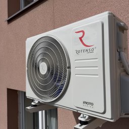 HYDROLINE SPÓŁKA Z OGRANICZONĄ ODPOWIEDZIALNOŚCIĄ - Biała jednostka zewnętrzna klimatyzacji Rotenso Imoto Series zamontowana na elewacji budynku, widoczna z boku, z oknem w tle. Jasne, naturalne oświetlenie.