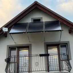 HYDROLINE SPÓŁKA Z OGRANICZONĄ ODPOWIEDZIALNOŚCIĄ - Trzy panele słoneczne zamontowane nad balkonem domu jednorodzinnego, widok z dołu, z anteną satelitarną po lewej stronie. Balustrada balkonu z ozdobnymi elementami.